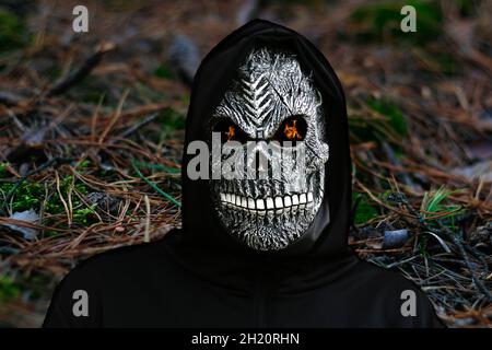 Nahaufnahme des grimmigen Reaper. Mann in Totenmaske mit Feuerflamme in den Augen auf dunklem, wildem Naturhintergrund. Faschingskostüm, gruselige Zähne. Halloween Stockfoto