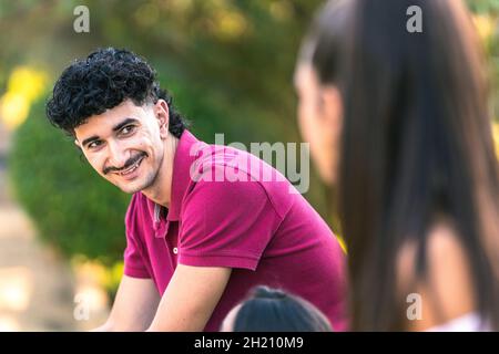 Mann mit moderner Frisur sitzt auf einer Bank, während er mit anderen Menschen spricht Stockfoto