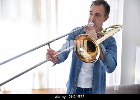Posaunist übt Posaune in einem Raum mit Vorhang im Hintergrund zu Hause. Horizontale Zusammensetzung. Stockfoto
