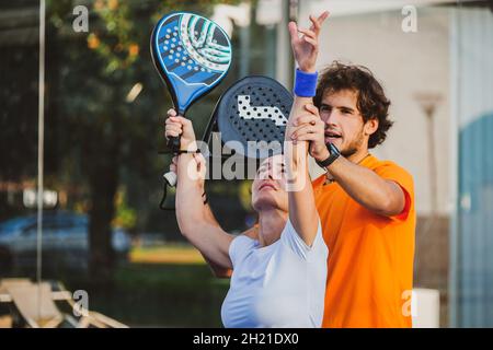 Der junge Lehrer überwacht den Padel-Unterricht für seine Schülerin - Coach lehrt Mädchen, wie man Padel auf dem Tennisplatz im Freien spielt Stockfoto