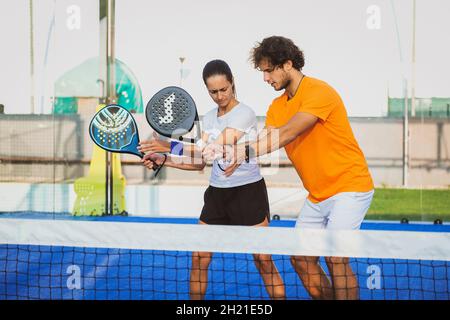 Der junge Lehrer überwacht den Padel-Unterricht für seine Schülerin - Coach lehrt Mädchen, wie man Padel auf dem Tennisplatz im Freien spielt Stockfoto