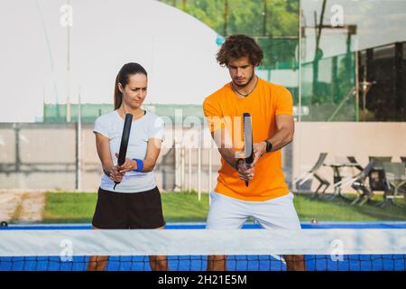 Der junge Lehrer überwacht den Padel-Unterricht für seine Schülerin - Coach lehrt Mädchen, wie man Padel auf dem Tennisplatz im Freien spielt Stockfoto