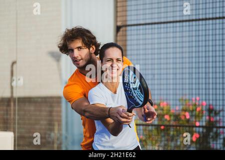 Der junge Lehrer überwacht den Padel-Unterricht für seine Schülerin - Coach lehrt Mädchen, wie man Padel auf dem Tennisplatz im Freien spielt Stockfoto