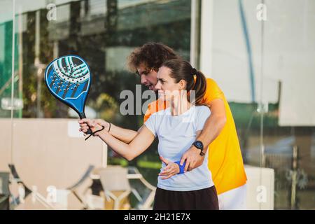 Der junge Lehrer überwacht den Padel-Unterricht für seine Schülerin - Coach lehrt Mädchen, wie man Padel auf dem Tennisplatz im Freien spielt Stockfoto