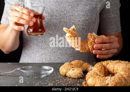 Köstlicher türkischer Bagel mit Sesamsamen, bekannt als Susamli simit. Eine Frau isst ihn zusammen mit schwarzem Tee, der in türkischen Teetassen serviert wird. Dunkle Rückseite Stockfoto