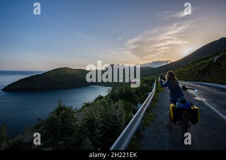 Die blonde kaukasische Radfahrerin ist unterwegs und macht ein Foto. Keem Bay, Achill Island, Irland Stockfoto