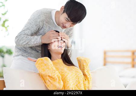 Lächelnder junger Mann, der die Augen seines Partners bedeckt. Junger Mann überraschend Frau im Wohnzimmer zu Hause. Stockfoto