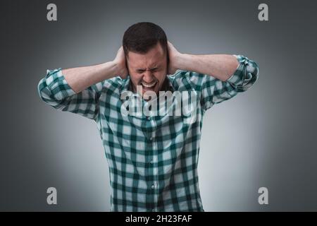 Frustrierter, gutaussehender Mann, der seine Ohren bedeckt und die Hände vor grauem Hintergrund stehen. Stockfoto