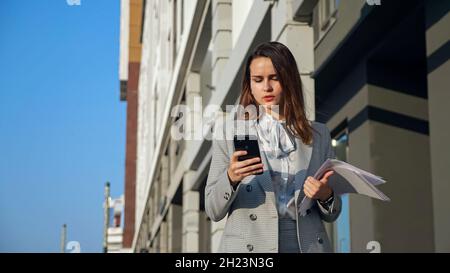 Junge Frau in einem Geschäftsanzug mit einem Telefon und Dokumenten, die die Straße entlang gehen Stockfoto