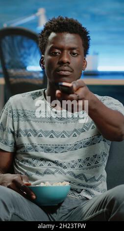 POV von Erwachsenen umschalten Kanäle mit TV-Fernbedienung. Person, die Fernsehen schaut und den Sender wechselt, während sie Popcorn-Snacks für die Freizeitunterhaltung hat. Mann, der die Kamera anschaut Stockfoto