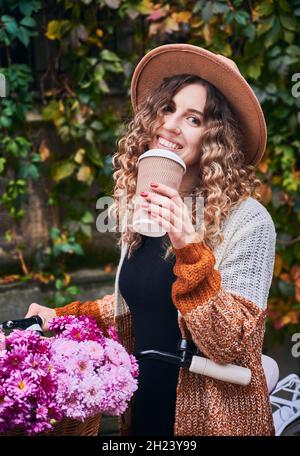 Schöne junge Frau im Hut hält eine Tasse Kaffee und lächelt, während sie neben dem Fahrrad mit Blumen im Korb steht. Stilvolle Frau mit Fahrrad trinken Kaffee, um im Freien zu gehen. Stockfoto