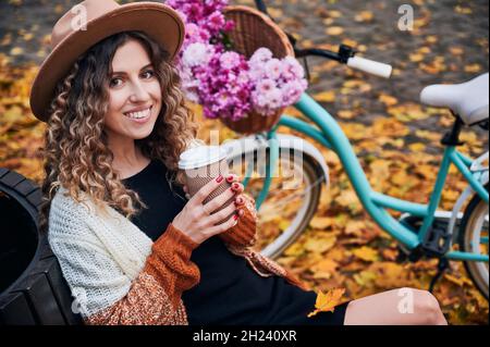 Portrait von attraktiven Mädchen mit lockigen Haaren, braune Augen. Frau, die sich auf der Bank ausruht und Kaffee trinkt. Auf dem Hintergrund städtischen blauen Fahrrad mit Korb von Blumen geparkt auf Bürgersteig mit gefallenen Blättern bedeckt. Stockfoto