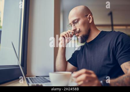 Fokussierter Hipster Millennial-Mann arbeitet am Laptop, während er im Café mit Kaffee sitzt. Freiberufliches Konzept Stockfoto