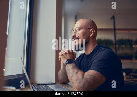 Der denkende Millennials-Hipster arbeitet am Laptop, während er im Café mit Kaffee sitzt. Freiberufliches Konzept Stockfoto