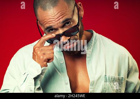 Porträt eines swarthy Latino fragenden Blick von unter Sonnenbrille auf einem roten Hintergrund. Brutaler bärtiger Mann mit einem kurzen Haarschnitt. Stockfoto
