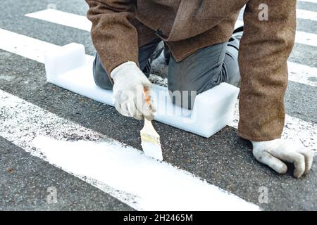 Ein Mann malt mit einem Pinsel auf die Asphaltmarkierungen für eine Fußgängerüberfahrt. Straßenreparatur. Stockfoto