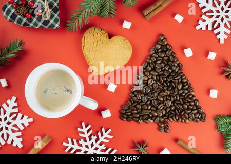 Braune Kaffeebohnen in Form eines Weihnachtsbaums mit Fichtenzweigen, einer Tasse Kaffee, einem herzförmigen Keks und hölzernen Schneeflocken auf rotem Backgroun Stockfoto