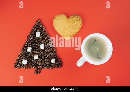 Braune Kaffeebohnen und Marschwalben in Form eines Weihnachtsbaums mit einer Tasse Kaffee und einem herzförmigen Keks auf rotem Hintergrund. Flach liegend Stockfoto