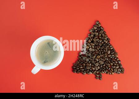 Braune Kaffeebohnen in Form eines Weihnachtsbaums mit einer Tasse Kaffee auf rotem Hintergrund. Flach liegend. Speicherplatz kopieren Stockfoto