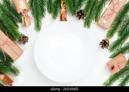 Weiße leere Keramikplatte mit Kopierfläche auf weißem Tisch mit Weihnachtsdekorationen aus Tannenzweigen, Zapfen und Geschenken Stockfoto