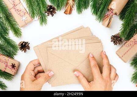 Flaches Lay von zwei weiblichen Händen, die Stapel von Bastelpapier-Umschlägen für Grußkarten halten, Weihnachtshintergrund von Tannenzweigen auf weißem Tisch Stockfoto