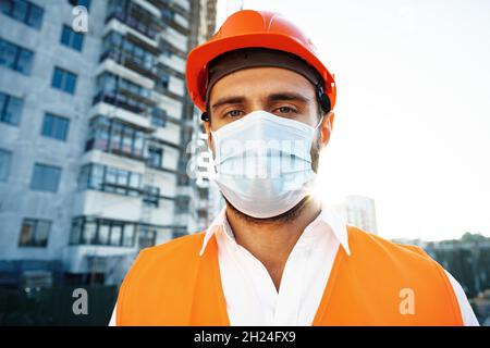 Porträt eines Baumeisters in Arbeitskleidung und Hardhat mit medizinischer Maske, Nahaufnahme Stockfoto