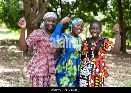 Eine Gruppe junger schwarzafrikanischer Dorfbewohner in farbenfrohen traditionellen Kleidern lächelt mit geballten Fäusten als Symbol für die Streng der Frauen an der Kamera an Stockfoto