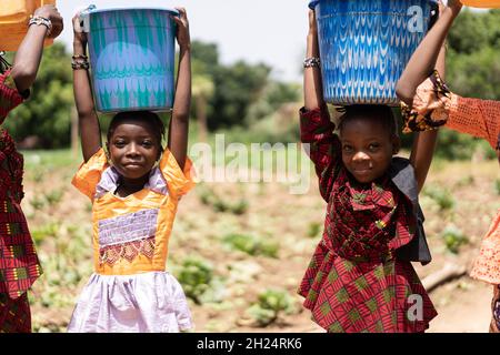 Nahaufnahme von zwei kleinen schwarzen afrikanischen Mädchen, die blaue Wassereimer auf ihren Köpfen trugen; Kinderarbeit Konzept Stockfoto