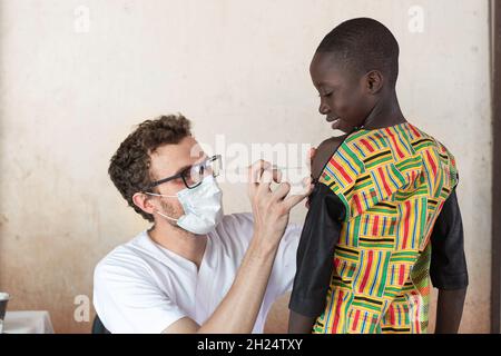 Gesundheitsprofesional trägt eine Gesichtsmaske sorgfältig Vorbereitung Malariaimpfung in einem kleinen schwarzen afrikanischen Schuljungen Stockfoto