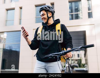 Lächelnder Lieferer, der mit dem Fahrrad unterwegs ist und in der Stadt auf das Handy schaut Stockfoto