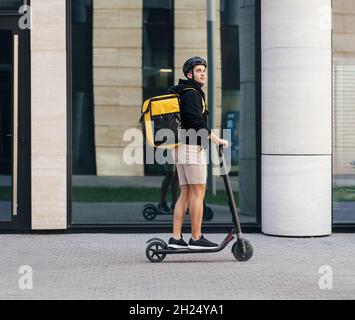 Seitenansicht des männlichen Kurierers, der auf einem elektrischen Push-Roller mit Rucksack für die Lebensmittelzustellung fährt Stockfoto