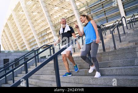 Sportliches Paar mittleren Alters, Mann und Frau in Sportkleidung diskutieren etwas, während sie die Treppe hinunter gehen, nachdem sie gemeinsam im Freien trainiert haben Stockfoto