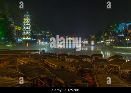 Die Wanming-Pagode und überdachte Boote auf dem Fluss Tuojiang in der Nacht in Fenghuang, China. Stockfoto