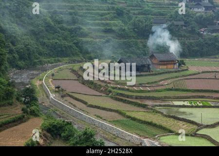 Rauch steigt aus einem Bauernhaus, das von terrassenförmigen Reisfeldern in der ländlichen Provinz Hunan, China, umgeben ist. Stockfoto