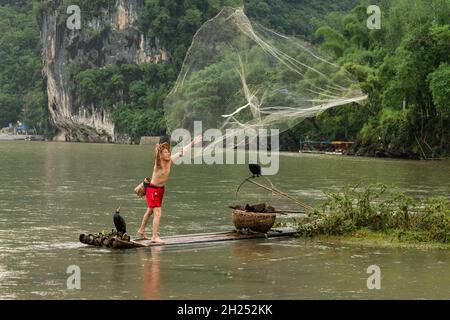 Ein Kormoran-Fischer in einem kegelförmigen Hut auf einem Bambusfloß wirft ein gegossenes Netz in den Fluss Li, Xingping, China. Stockfoto