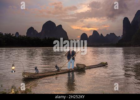 Kormoran Fischer in konischem Hut & Umhang auf Bambusfloß mit Kormoranen & Laterne. Li River, China. Stockfoto