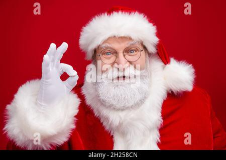 Foto von lustig aufgeregt pensionierten Mann tragen weihnachtsmann Kostüm Brille lächelnd zeigt okey Zeichen isoliert roten Hintergrund Stockfoto