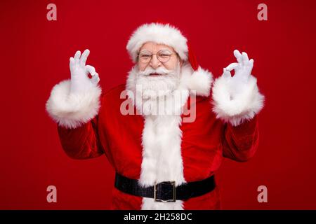 Foto von selbstbewussten gut aussehenden Mann Rentner gekleidet weihnachtsmann Kostüm zeigt okey Zeichen lächelnd isoliert roten Hintergrund Stockfoto