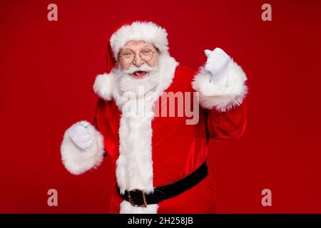 Foto von beeindruckt glücklichen Mann Rentner gekleidet weihnachtsmann Kostüm aufsteigende Fäuste lächeln isoliert roten Hintergrund Stockfoto