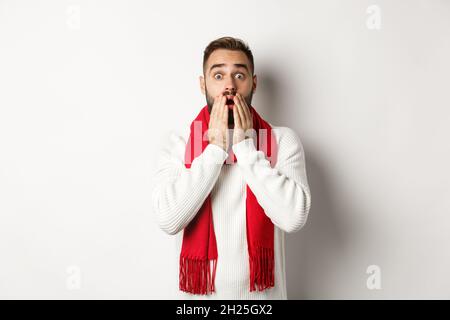 Weihnachtsferien und Feier Konzept. Schöner bärtiger Kerl in rotem Schal und Pullover, der schockiert, beeindruckt von Werbung, weiß Stockfoto