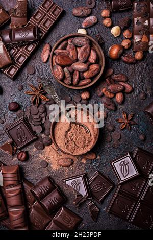 Stücke gebrochener leckerer Milch und dunkler Schokolade mit Kakaobohnen, Haselnuss und Pulver auf schwarzem Beton-Hintergrund Stockfoto