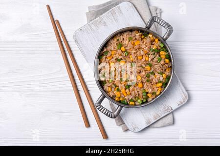 Draufsicht auf Bratpfanne mit chinesischem Reis, Hühnereiern, gelbem Mais und grünen Zwiebeln auf Holzbrett und weißem Holz Hintergrund Stockfoto