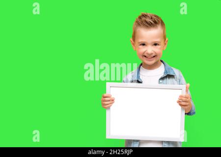 Der fünfjährige Junge lächelt und hält einen weißen Rahmen. Glücklich Vorschulkinder isoliert auf grünem Hintergrund mit Platz für die Kopie der Nachricht, Informationen, m Stockfoto