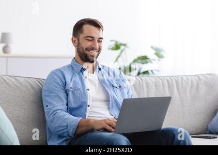 Lächelnder Kaukasier mittleren Alters mit Bart sitzt auf dem Sofa und arbeitet im Wohnzimmer auf einem Laptop Stockfoto