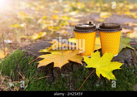 Nahaufnahme von zwei Pappkaffeebechern, die auf einem Baumstumpf stehen, der mit Moos und Herbstblättern bedeckt ist, vor einem verschwommenen Herbstlaub. Stockfoto