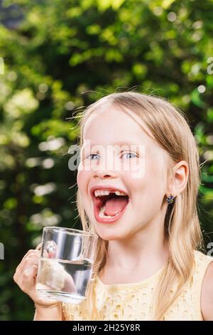 Glückliches kleines Mädchen trägt gelbes Kleid hält Glas Tasse Wasser in einem Sommergarten - gesunde Lebensweise Konzept Stockfoto