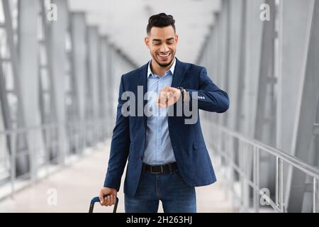 Junger Arabischer Geschäftsmann, Der Mit Dem Koffer Im Flughafen Unterwegs Ist Und Die Armbanduhr Anschaut Stockfoto