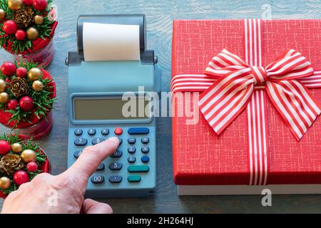 Eine Hand, die die Knöpfe an der Kasse neben einer roten Geschenkbox mit Band und kleinen Weihnachtsbäumen mit Zapfen drückt. Berechnung des Betrags Stockfoto