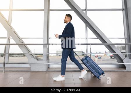 Junger Geschäftsmann aus dem Nahen Osten mit Koffer und Smartphone in den Händen, der auf dem Flughafen unterwegs ist Stockfoto