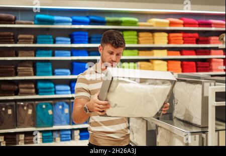 Der Mann las Informationen über einen Kasten mit Bettwäsche, während er in einem modernen Einkaufszentrum mit farbenfrohen Handtüchern und Bademänteln gegen die Regale stand Stockfoto
